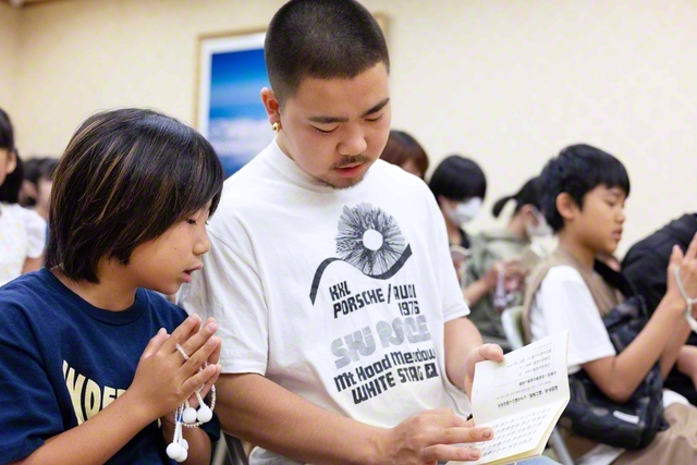 高校3年のノエルさん㊥が、経本の文字を指で丁寧になぞる。それを小学2年の里乃さん㊧が目で追いながら、勤行を元気に行う。会場には、この2人のように勤行を一緒にする姿がほかにも見られた