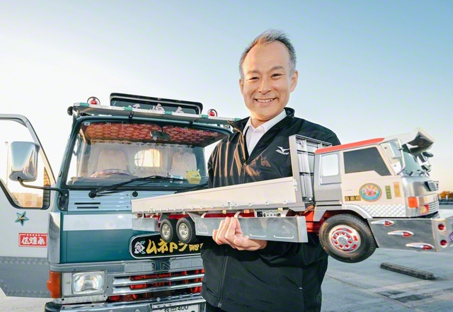 腕に抱いた自作のデコトラ模型は、操縦も可能。「カッコイイし、カワイイでしょう。しかも、これを待ってくれる仲間がいる。なんて楽しい人生でしょうか。信心と一緒。好きだから言いたくなっちゃうんですよ」と宗藤さん。写真後方は自家用車。もちろんデコトラである