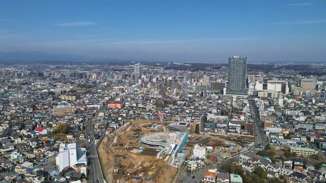 かつてシュリーマンが八王子に入る時に歩いたであろう現・子安町の上空から、北の方角を望む。右のタワーマンションが立つ地点がJR八王子駅周辺。中央のかなたには、丹木の丘に立つ創価大学の本部棟がかすかに見える。手前の工事中のエリアには本年秋、公園や図書館、ミュージアム、交流スペースが一体となった拠点「桑都（そうと）の杜（もり）」が誕生する