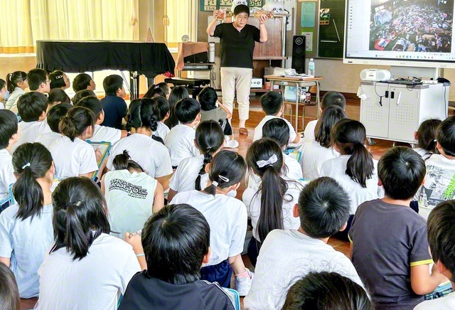 小学校で語り部を行う鎌田さん（奥）。菓子パンを手に「同じ物を食べ続けるのは苦しかった。でも、この一つのパンが命をつないでくれた」と訴える（医療ネットワーク支援センター提供）