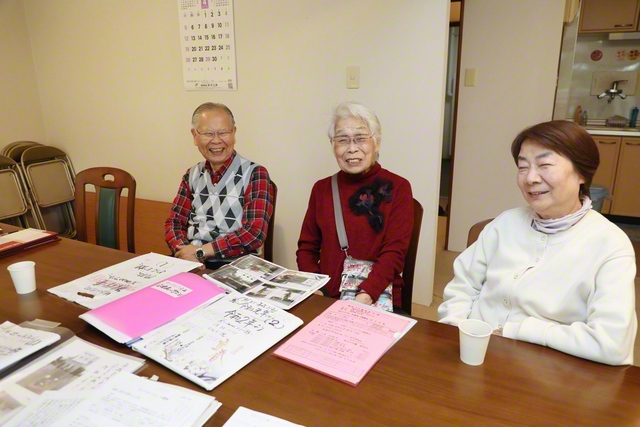 満生さん㊥と田中さん夫妻が和やかに。机には、「ふれあいサロン」や地域活動での思い出の品が