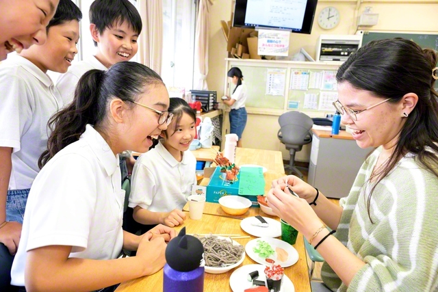 紙や毛糸で作った食べ物が並ぶ手作り屋台を囲み、笑顔で交流する児童と留学生（東京創価小学校で）