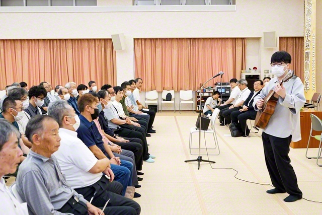江口純一さんの司会でスタートした飯塚東本部の集い。金子秀樹さんがギター演奏を披露し、会場を盛り上げた（21日、飯塚平和会館で）