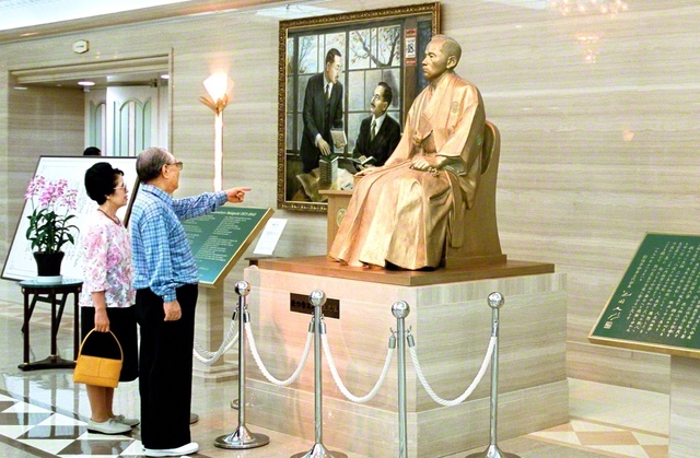 牧口先生の座像を見つめる池田先生ご夫妻（2001年7月、東京牧口記念会館で）。池田先生は牧口先生の生き方を通し、「青年とは、決して年齢ではない。心の持ち方、生き方で決まる」と