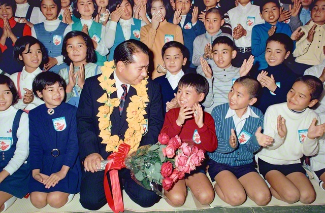 首に掛けられた真心のレイ、手元には鮮やかな赤い花束――。1972年1月30日、コザ市（現・沖縄市）の諸見会館で行われた記念撮影会で、池田先生が未来部の友を温かく励ました