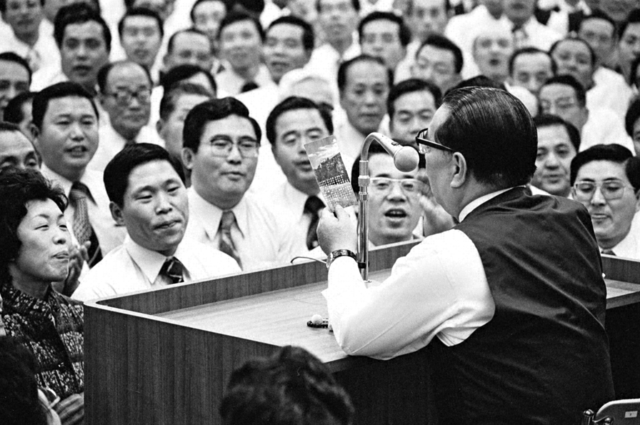 同志が師の元へ駆け付けて開催された県支部長会。池田先生が「学会歌を歌おう！」と提案し、皆で県歌「広布の旗」を大合唱した。先生の手には、最前列の友が手渡した歌詞カードが（1979年11月、神奈川文化会館で）
