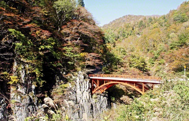 【津南町　前倉橋と布岩山】中津川渓谷に架かる赤いアーチが目を引く前倉橋。その近くには柱状節理の絶壁である布岩山がそびえ立つ。岩肌からのぞく赤や黄色の紅葉のコントラストが美しい。秋本番、「全山紅葉の勝利山」へ、題目根本に対話拡大の輪を広げてまいります（23日撮影、小島文雄通信員）
