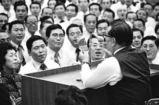 同志が師の元へ駆け付けて開催された県支部長会。池田先生が「学会歌を歌おう！」と提案し、皆で県歌「広布の旗」を大合唱した。先生の手には、最前列の友が手渡した歌詞カードが（1979年11月、神奈川文化会館で）