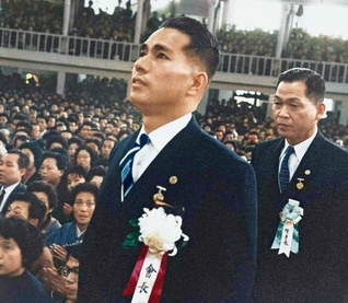 第3代会長就任式で入場する池田先生。その視線の先には恩師・戸田先生の遺影が掲げられていた（1960年5月3日、東京・日大講堂）。モノクロ（白黒）写真を、編集部の責任のもと、AI（人工知能）を活用してカラー化したもの