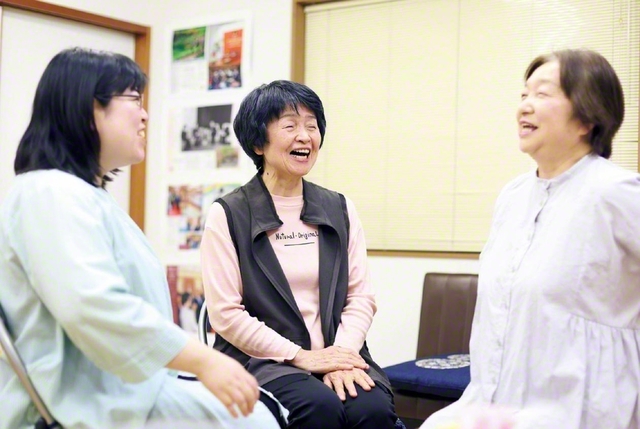自宅横にある地域の宝城で同志と語らう阿南さん㊥。この会場は地元の自治会の会合や子どもたちのイベント等でも使用されている。阿南さんは教育部のリーダーとしても活躍。孫の面倒も見ながら多忙な日々を送る。今回の「あしおと」掲載は、「夫が『これからも一緒に頑張るよ』と呼びかけてくれているのだと思います」と。今も心の中で夫妻二人三脚の前進を続けている