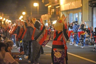 夜の町を進む踊り手。編みがさの下から伸びる手が、ちょうちんの明かりに浮かび上がる。奥へ続く踊りの列からも、祭りの熱気が伝わってくる