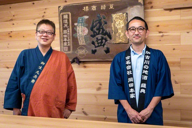 「合名会社岡田本家」6代目の岡田洋一さん㊨と看板の前で